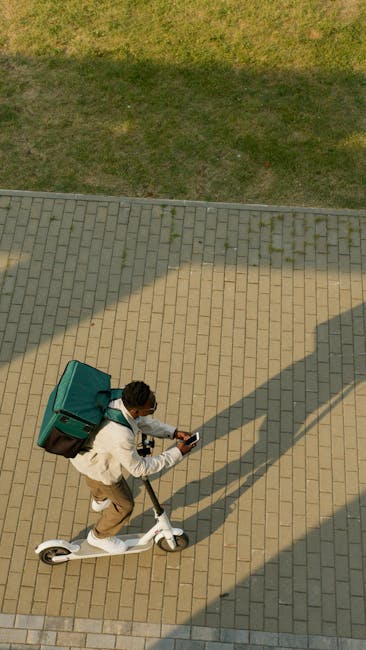 A person riding a white electric scooter on a paved brick pathway, carrying a large green delivery box on their back. The rider is wearing a white jacket, beige trousers, and has short dark hair. The