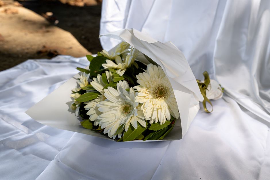 A bouquet of fresh white Gerbera daisies with large, open petals and prominent central disks, arranged with green foliage. The flowers are wrapped in elegant white tissue paper, secured with a gold ri