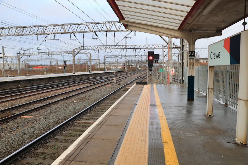 The image depicts Beckenham Junction train station platform BR3 on a cloudy day, with a wet, gray concrete surface and a yellow tactile strip along the edge. Several railway tracks extend into the dis