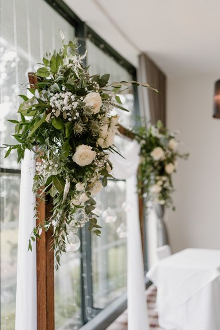 A pair of vertical wooden wedding arch decorations positioned indoors by large windows, each adorned with lush floral arrangements featuring white roses, baby’s breath, and various green foliage. Th