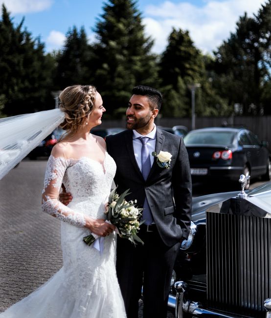 A bride and groom stand together outdoors on a paved surface, with a row of parked cars and lush green trees in the background under a partly cloudy sky. The bride, wearing a white lace wedding gown w